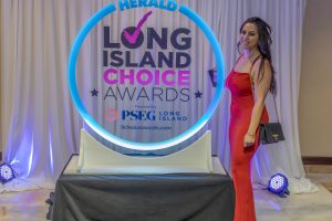 Woman in a red evening gown posing beside a glowing circular frame at the Long Island Choice Awards backdrop.