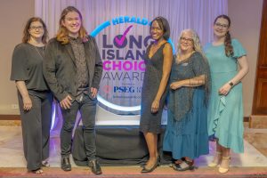 Five adults in formal attire pose on a stage in front of a blue and pink ‘Long Island Choice Awards’ backdrop at an event venue.