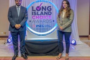 Two professionally dressed individuals stand on either side of a glowing circular backdrop reading Herald Long Island Choice Awards at an awards event.