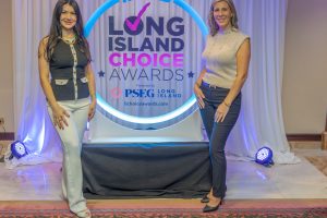 Two women pose on a prize‑ceremony stage in front of a 'Long Island Choice Awards' backdrop with a glowing circular sign above them.