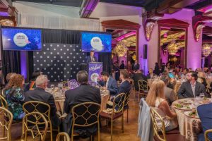 Speaker at podium on stage with Welcome sign; two screens show Long Island Choice awards, audience seated at round tables enjoying dinner in a glittering ballroom.