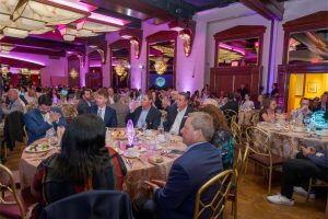 Large banquet hall full of guests in formal attire dining at round tables with pink-purple lighting and chandeliers in the background.