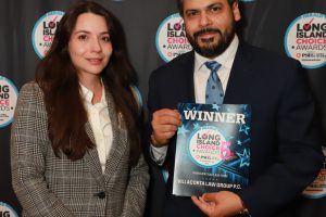 Two well-dressed individuals pose at an awards backdrop; the man on the right holds a 'WINNER' plaque for Long Island Choice Awards.