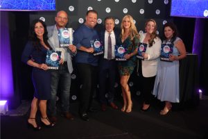 Eight people stand together on stage, posing with blue winner plaques at an awards ceremony, smiling at the camera.