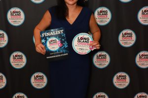 Woman in a blue dress stands in front of a Long Island Choice Awards backdrop, holding a finalist booklet and a round award badge, smiling at the camera.